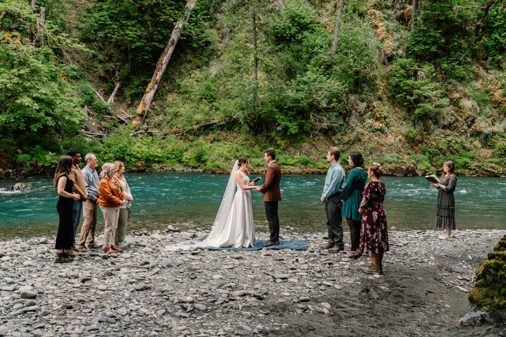 a wide shot of the ceremony from this backpacking wedding. a bright, blue river rushes behind our couple as they exchange vows, surrounded by their guests 