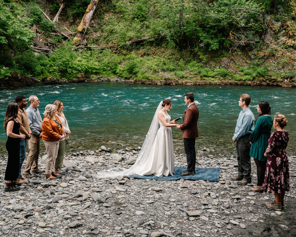 a wide shot of the ceremony from this backpacking wedding. a bright, blue river rushes behind our couple as they exchange vows, surrounded by their guests 