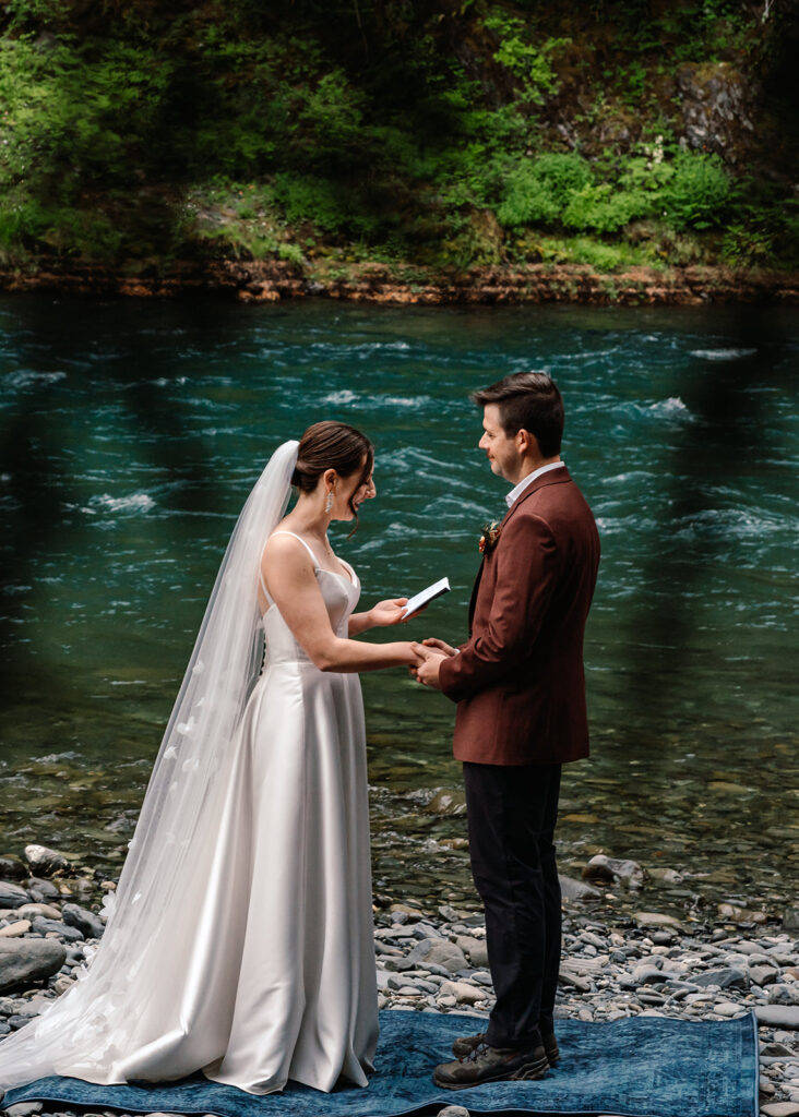 a bride reads her vows to her groom during their backpacking wedding. Behind them is a bright blue river. from the front, they are framed by pine leaves