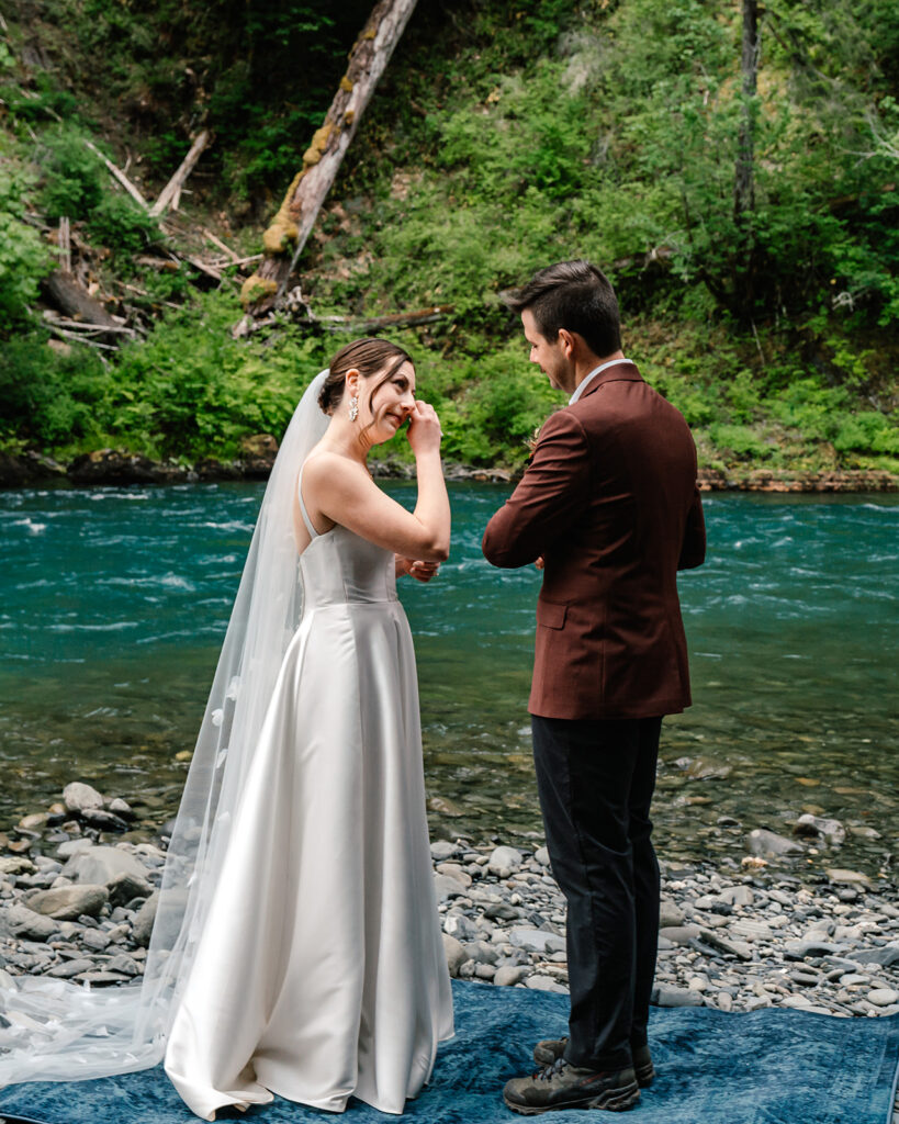 a bride wipes her eyes as her groom reads his vows. behind them is a gorgeous blue river, setting the scene for their ceremony during this backpacking wedding