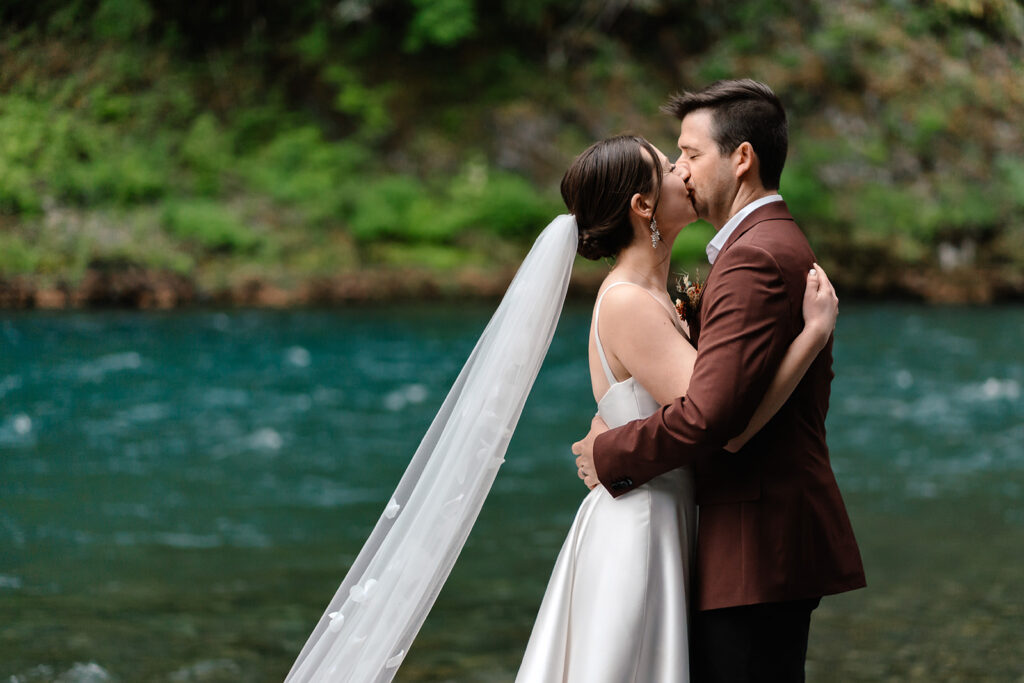 a bride and groom kiss in front of a bright, blue river during their backpacking wedding