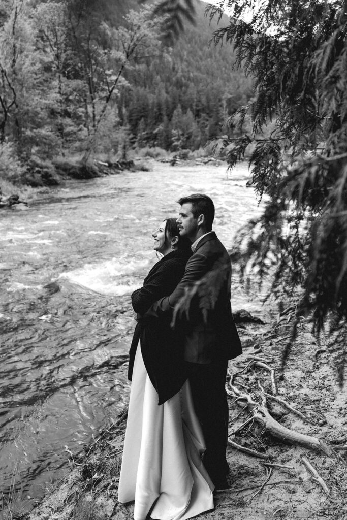 during their backpacking wedding, a bride and groom nestle up by the riverbed. She is wrapped in a dark blanket as her groom embraces her from behind