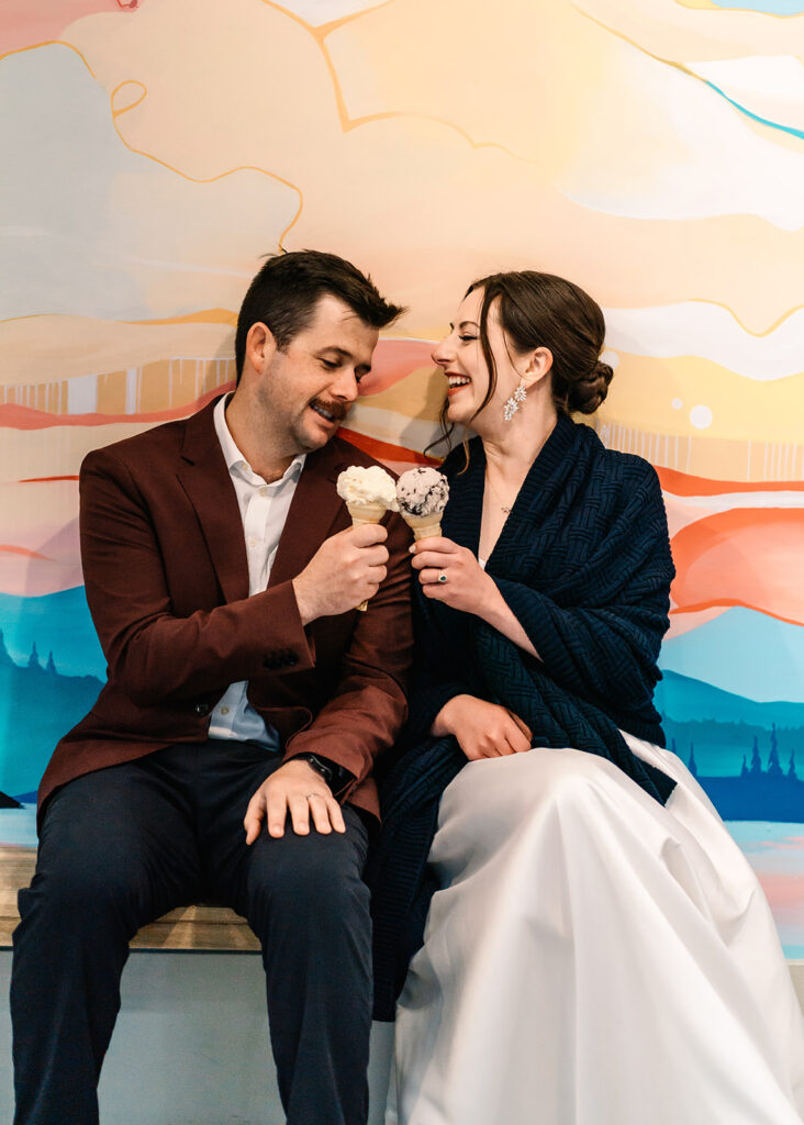 a bride and groom sit in front of a wall painted with bright colors. they cheers their cones as a way to end their backpacking wedding 