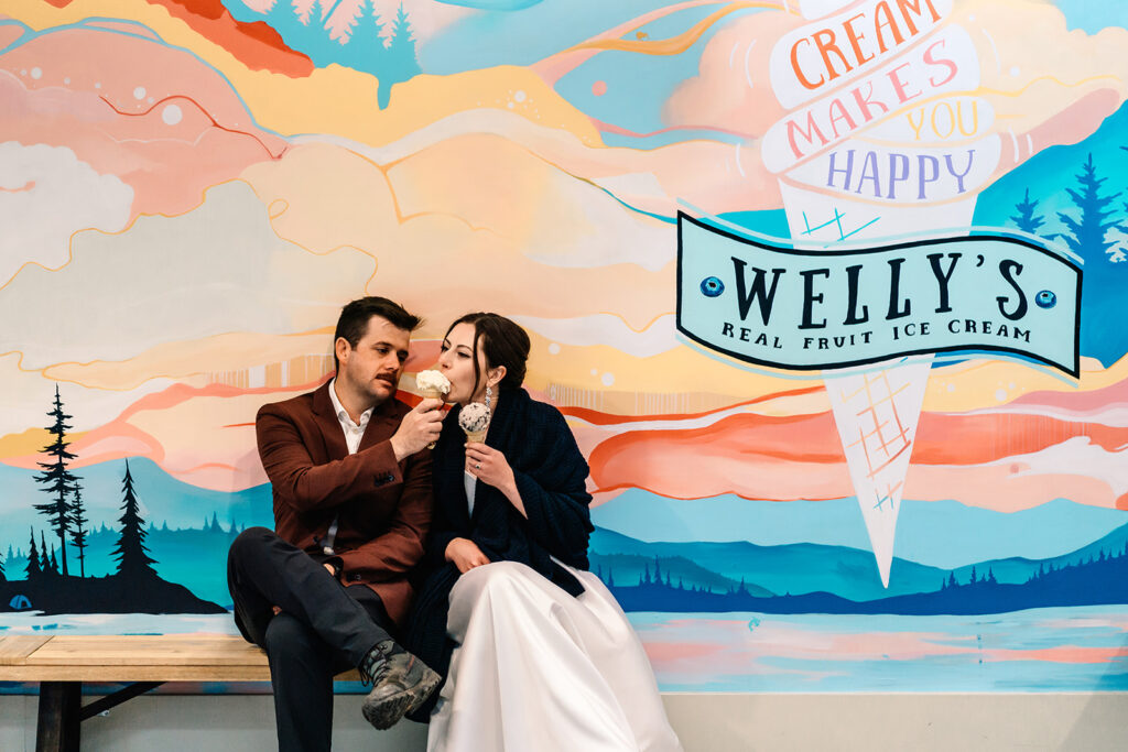 after their backpacking wedding, a bride and groom sit in front of a wall painted with bright colors. an ice bream cone is painted and reads "ice cream makes you happy". The bride takes a bite of her grooms ice cream cone 