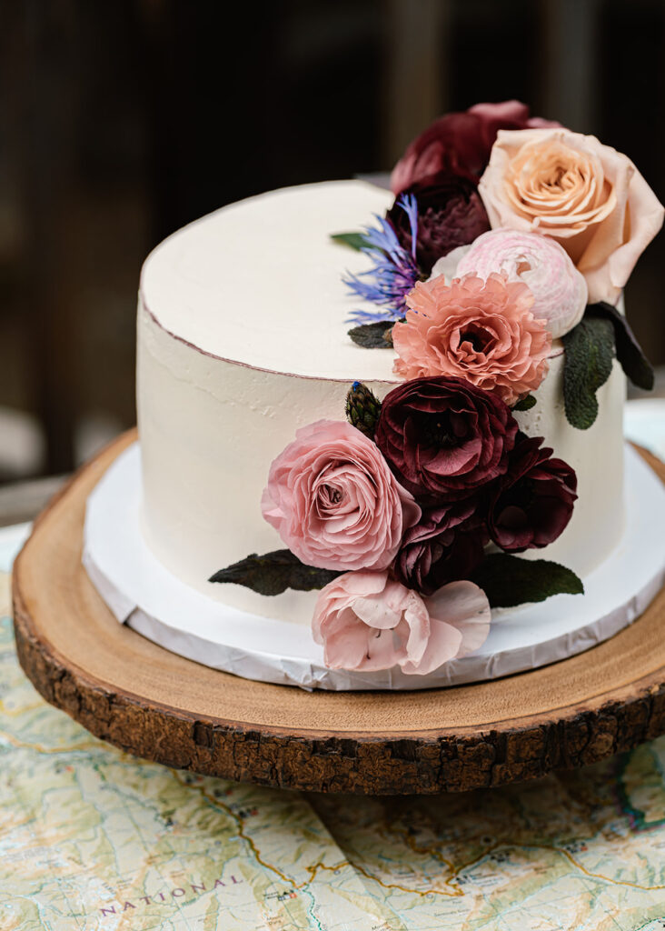 a vertical image of a single-tiered wedding cake with white frosting and a floral arrangement for this backpacking wedding 