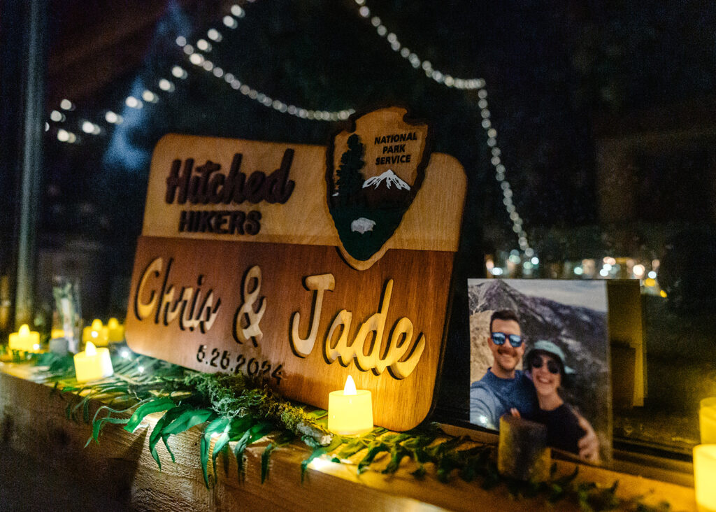 a national park style wooden sign reading "hitched hikers" personalized with their names and their wedding date is illuminated by candles and twinkle lights