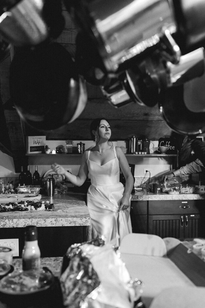 a black and white image of a bride dancing in the kitchen after her backpacking wedding. She is framed by hanging pots and pans