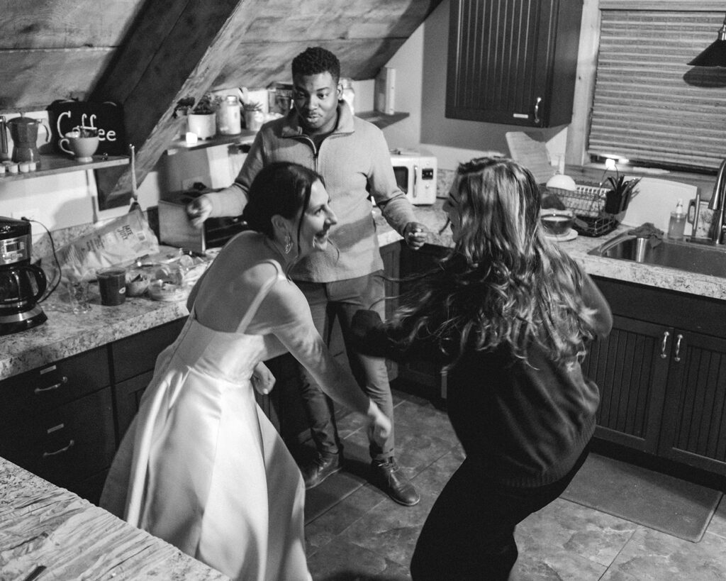 a black and white image of a bride dancing with her friends in the kitchen after her backpacking wedding