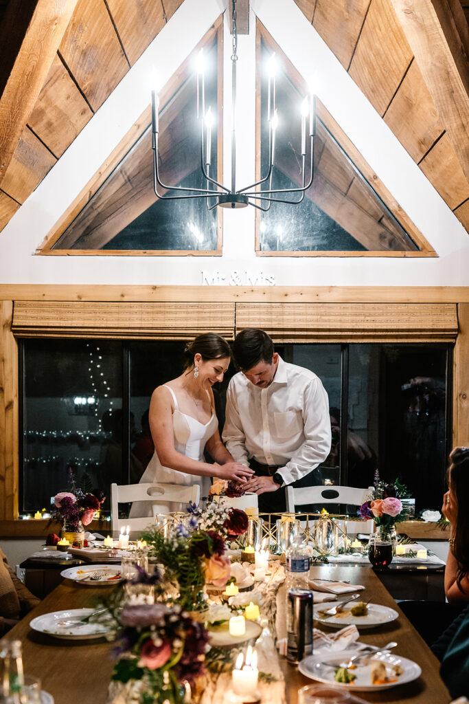 a bride and groom cut their wedding cake at a decorated table. they are framed by the structure of their a-frame cabin 