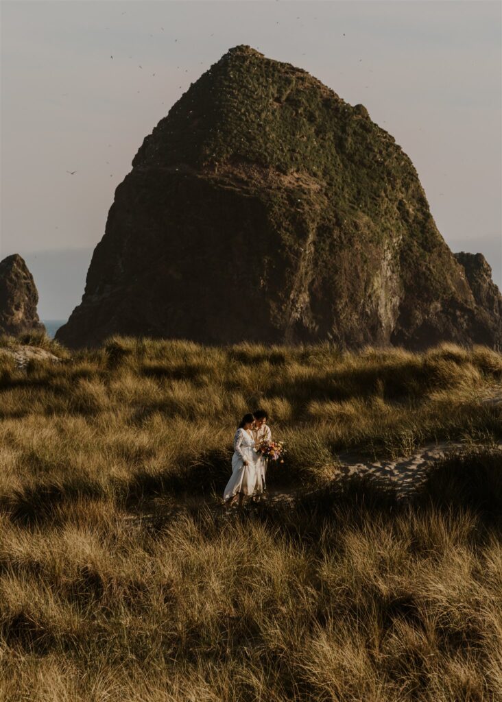 a bride and groom prepare for their elopement ceremony walking through tall sea grass that dances with the wind. Haystack rock frames their background. it is sunset and the light is gold 