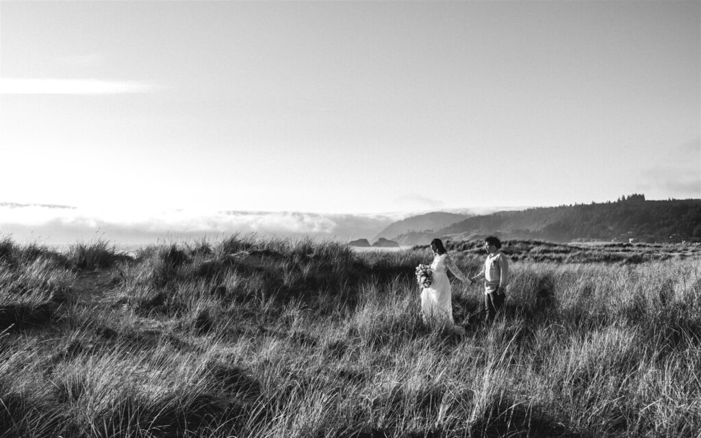 a black and white image of a bride leads her groom to their ceremony elopement location. Thy hold hands as she leads him through the tall sea grass