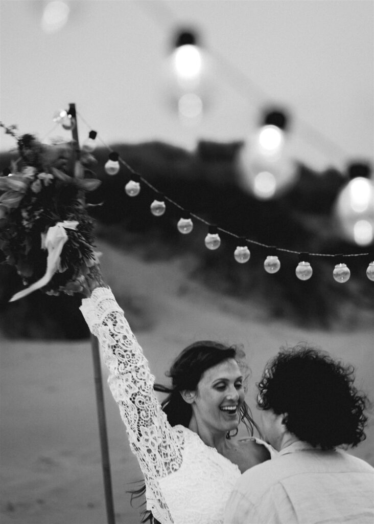 a black and white image of a bride and groom dancing under string lights to celebrate their elopement ceremony