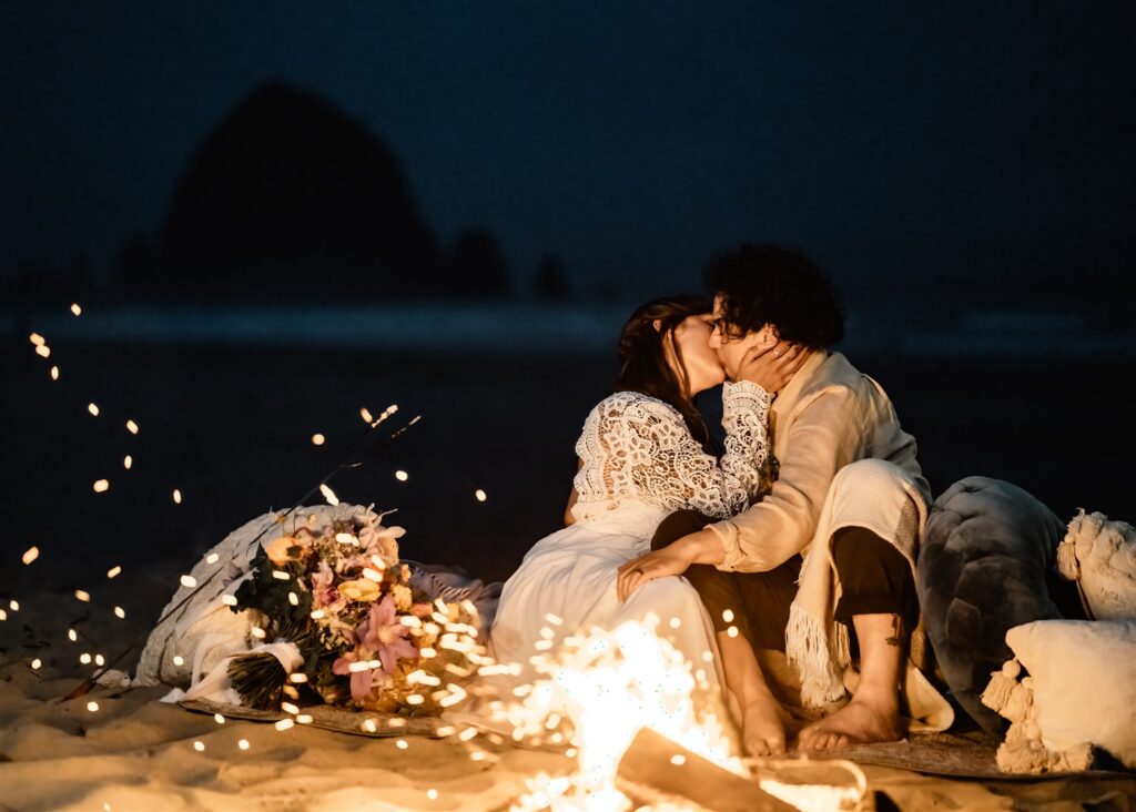 a bride and groom sit by a beach bonfire. they kiss as embers fly, celebrating after their elopement ceremony 