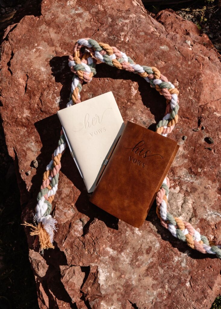 a shot of elopement ceremony details in harsh light on a marbled red rock. brown and white leather vow books and hand fasting cords 