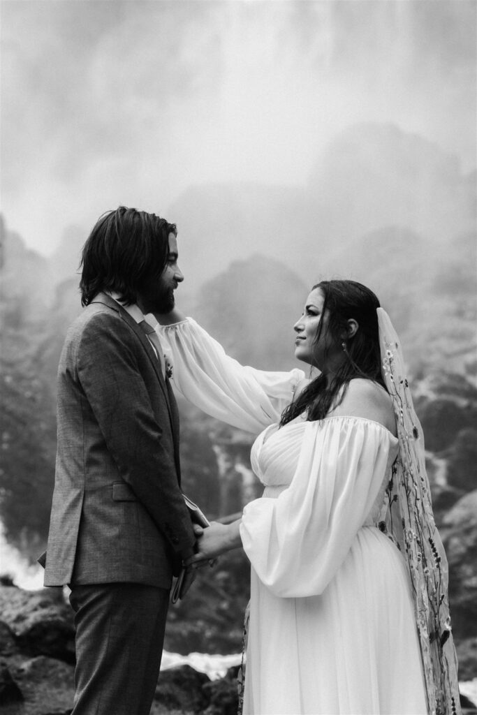 a bride tenderly caresses her grooms face during their elopement ceremony as a waterfall gushes behind them 