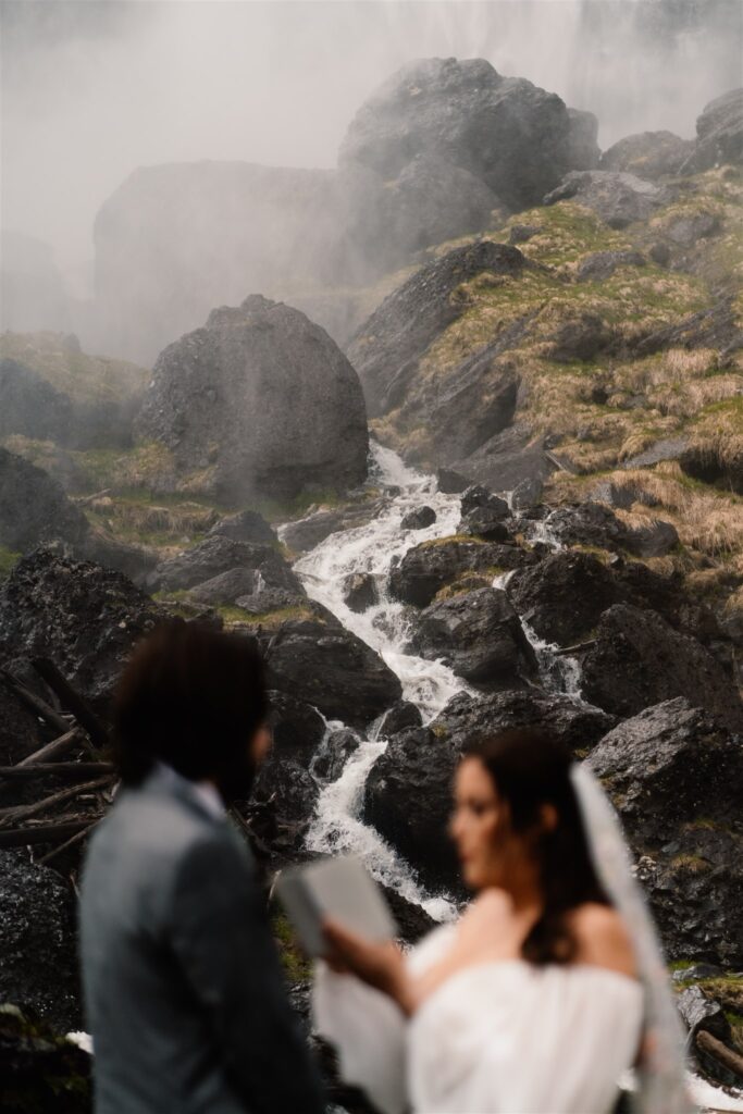 a small waterfall zig zags between a bride and groom. They frame the flowing pattern as she reads her vows during their elopement ceremony 