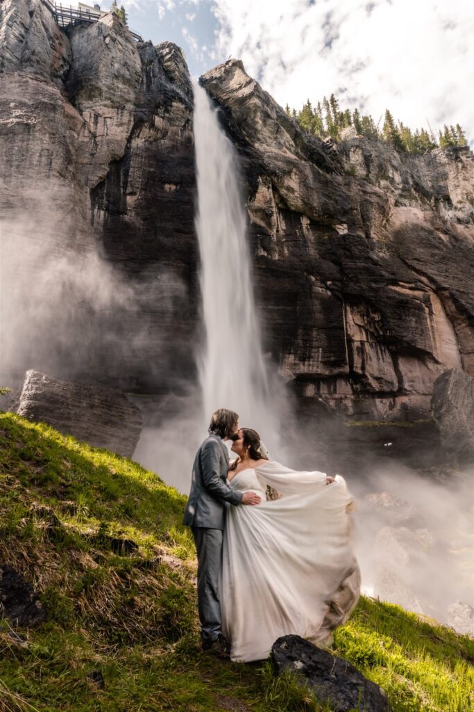 a groom kisses his bride on the forehead as here dress billows in the wind. a waterfall gushes behind them as they embrace during their elopement ceremony 