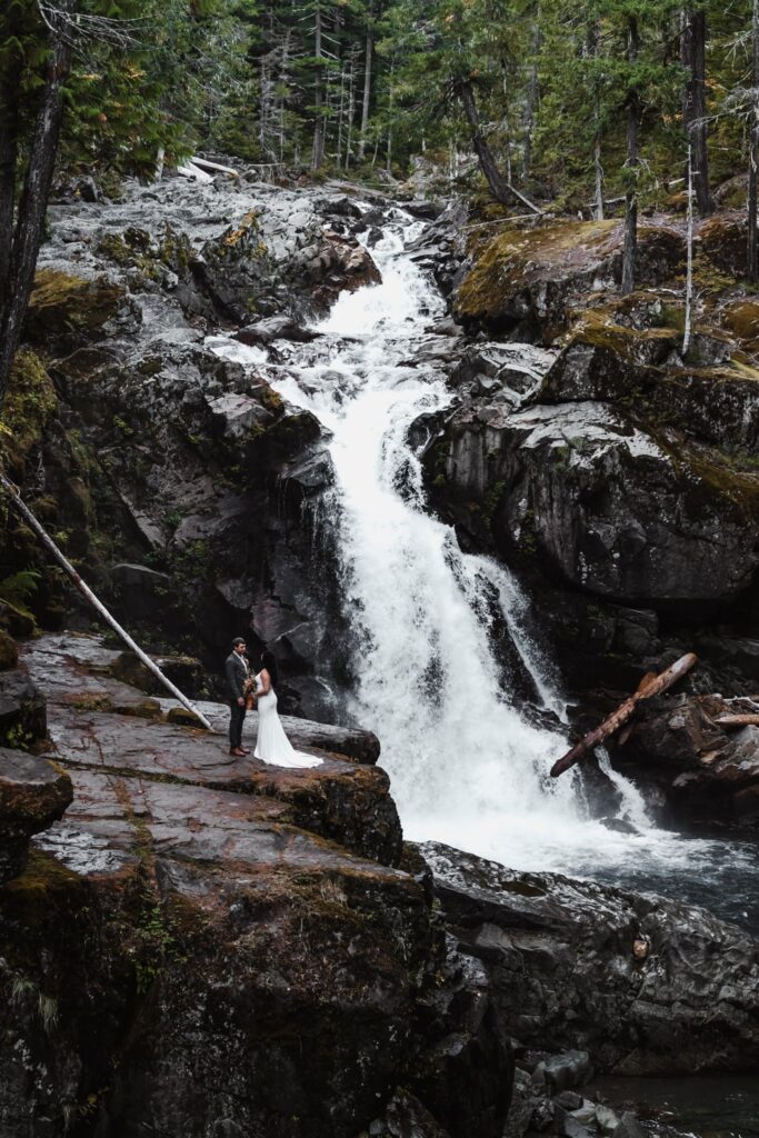  a bride and groom exchange vows during their elopement ceremony. They stand on the ledge of a cliff as a waterfall gushes behind them.