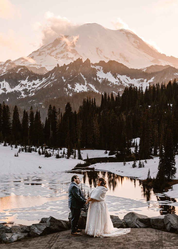 a bride and groom hold hands during their elopement ceremony. The sun sets behind a snowy mount Rainier 