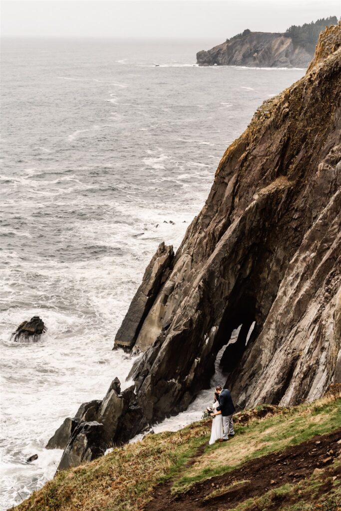 a bride and groom embrace while standing on a grassy cliff. Jagged sea cliffs make up their background