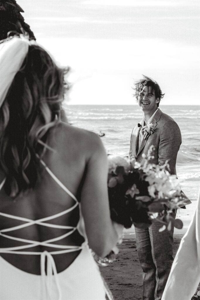 a black and white image of a groom smiling widely, his hair blowing in the wind. he is framed by the back of his bride as this shot was taken over her shoulder 