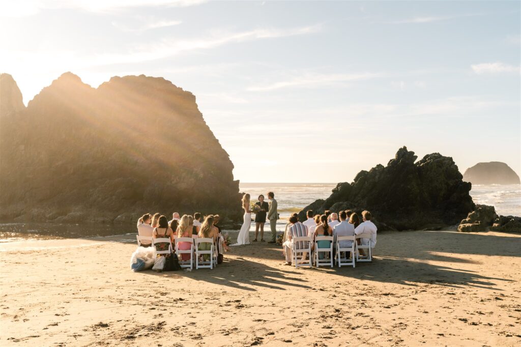 A landscape image of a beach elopement ceremony. Guest sit in chairs, watching a bride and groom exchange vows between two large sea stacks. 