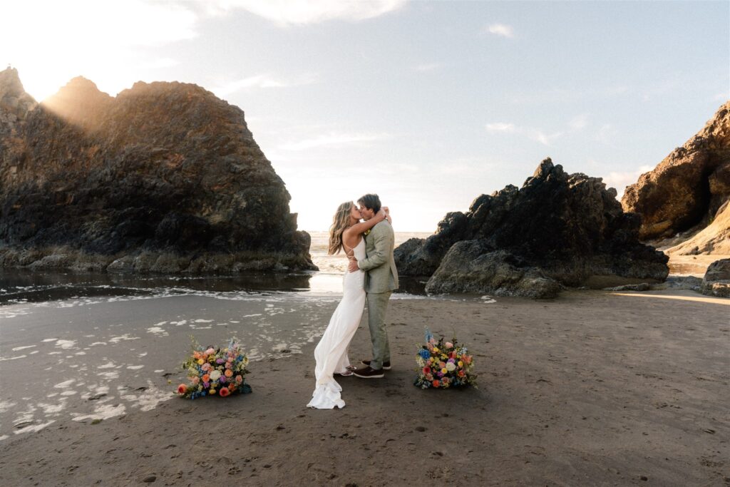 after their elopement ceremony, a bride and groom kiss between two large sea stacks. Small, brightly colored floral arrangements are at their feet as they embrace. 