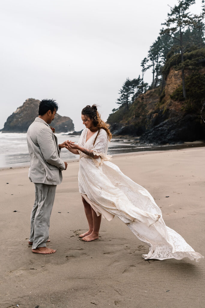 a bride places a ring on her grooms finger during their elopement ceremony on a moody beach. the wind whips her lace dress. they both stand barefoot on the sand