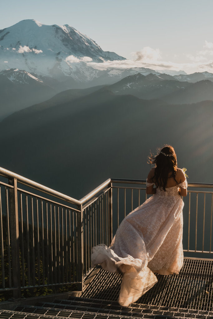 a bride gazes out at the blue mountains as her dress billows in the wind. The light is rose-gold as she looks out at Mount Rainier 