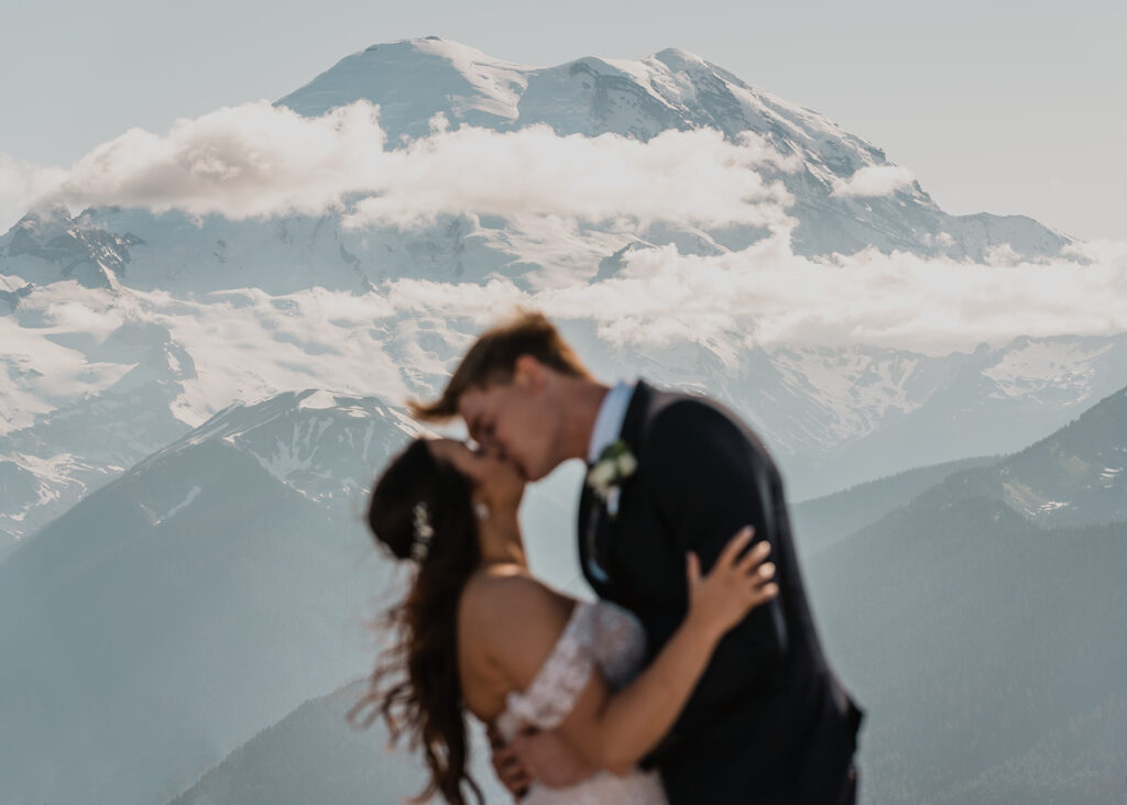 a bride and groom share their first kiss during their elopement ceremony. Mount Rainier is perfectly in focus as their bodies, slightly out of focus frame the landscape 