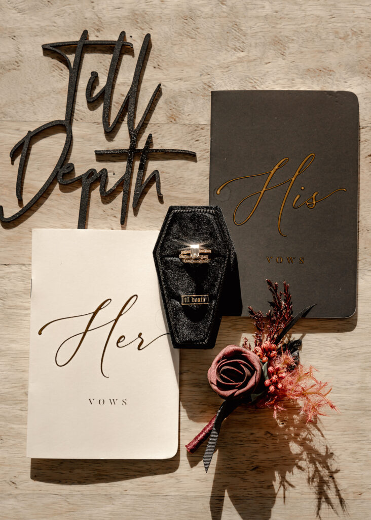 a shot of elopement ceremony details in harsh light. black and white vow books, a coffin wedding ring box, and a wine colored boutonnière 