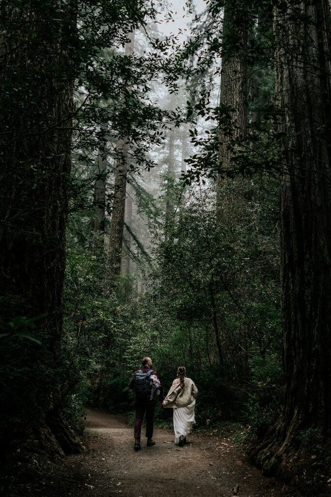 a bride and groom walk through a deeply wooded forest. they wear sweaters over their wedding attire as they journey to their elopement ceremony location