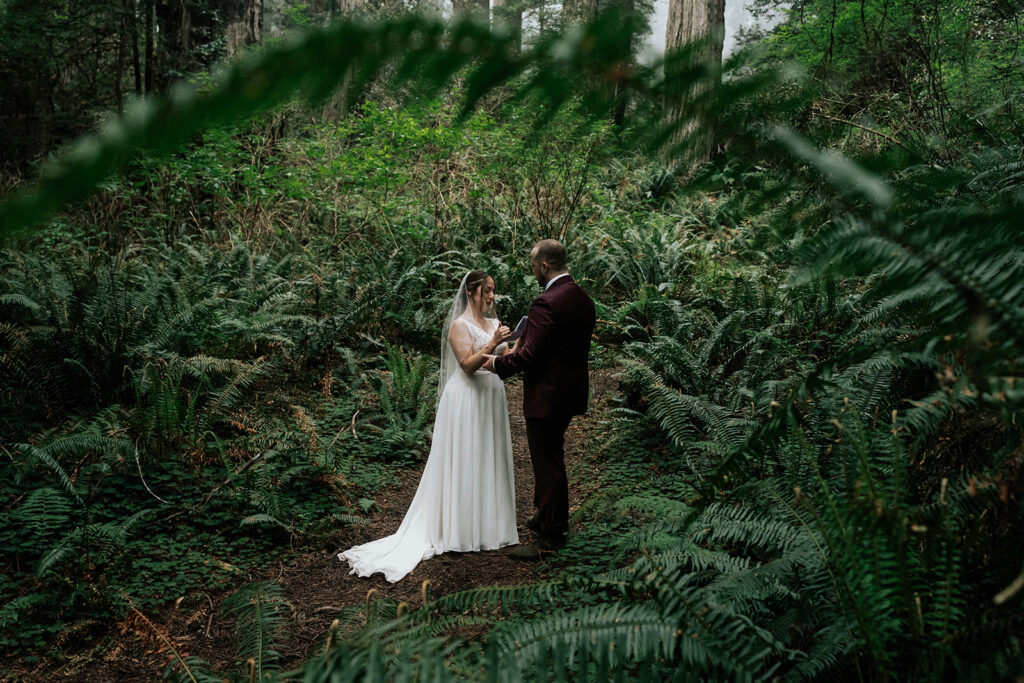 a bride reads her vows to her groom during their elopement ceremony. They stand in a forest framed by lush, green ferns