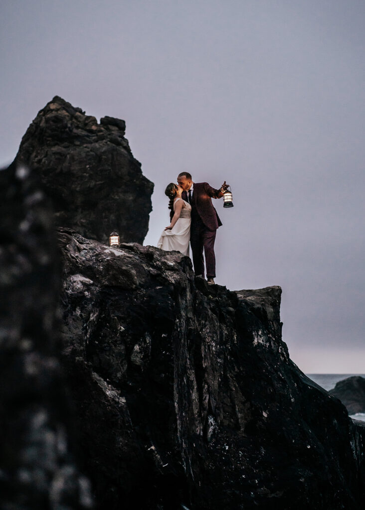 a bride and groom kiss at the top of a large sea rock during their elopement ceremony. It is just after sunset, giving us a pale, blue light. They hold lanterns to illuminate the landscape around them 