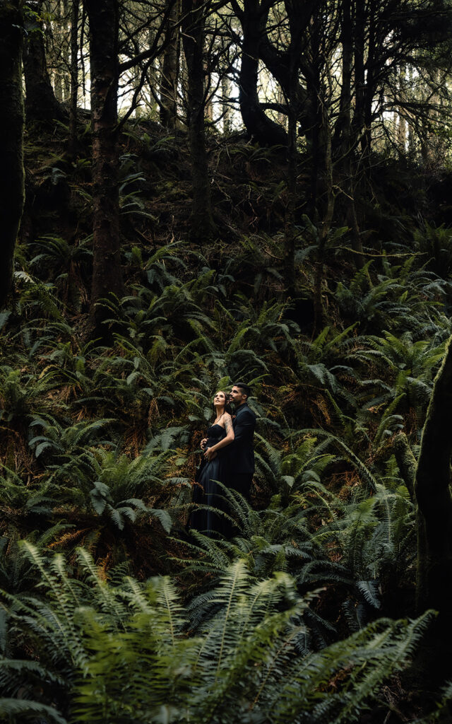 a bride and groom in all black wedding attire embrace in a lush, green forest. they look towards the canopy as the soft light shines on their faces. 