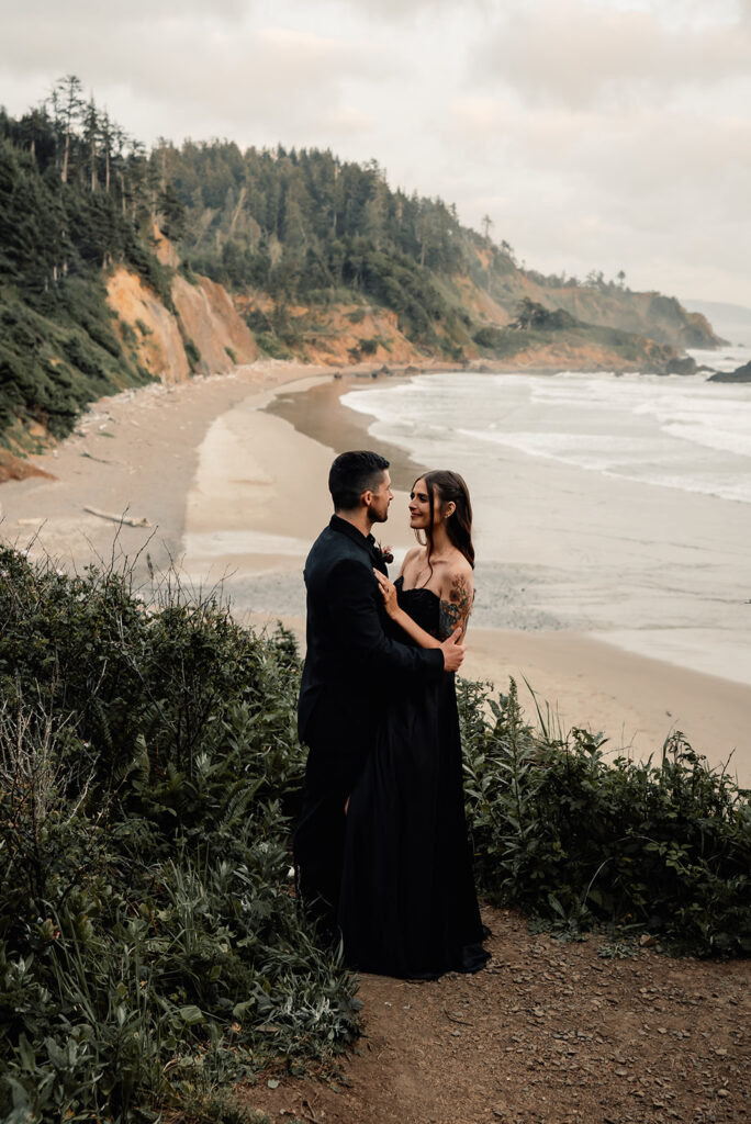 a bride and groom in all black wedding attire embrace during their elopement ceremony. the sea is seen below them as they stand on a cliff gazing out 