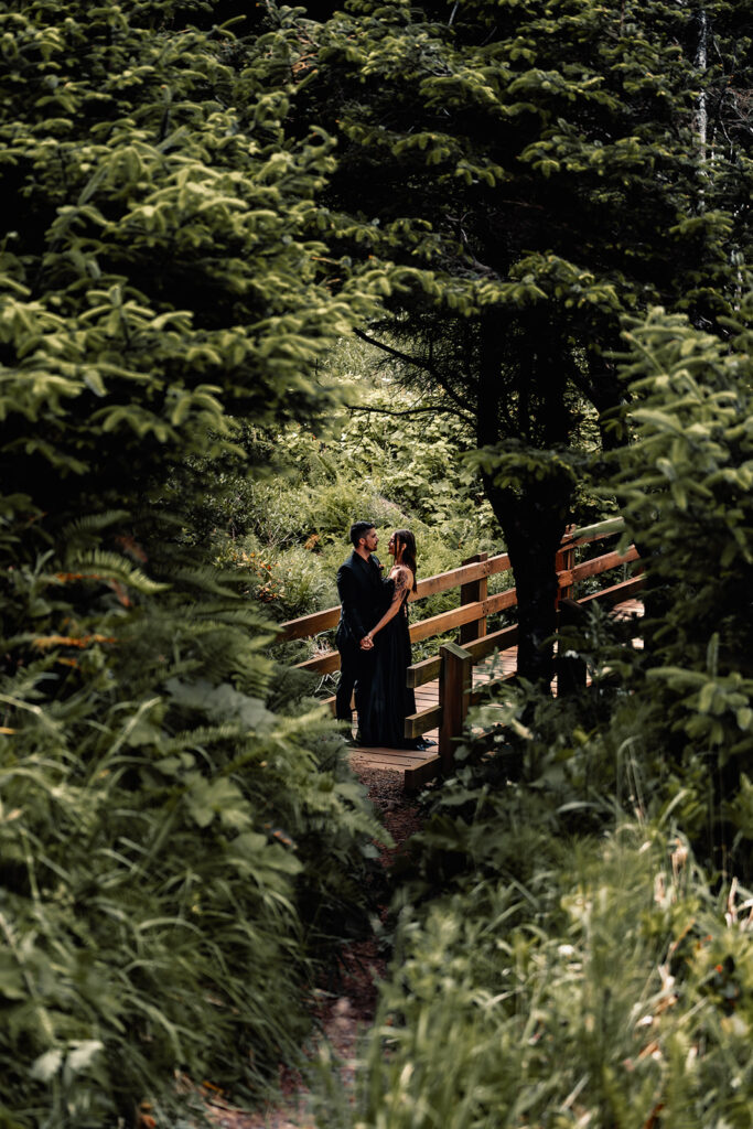 a bride and groom embrace on a wooden bridge. They wear all black wedding attire and are framed by green leaves 