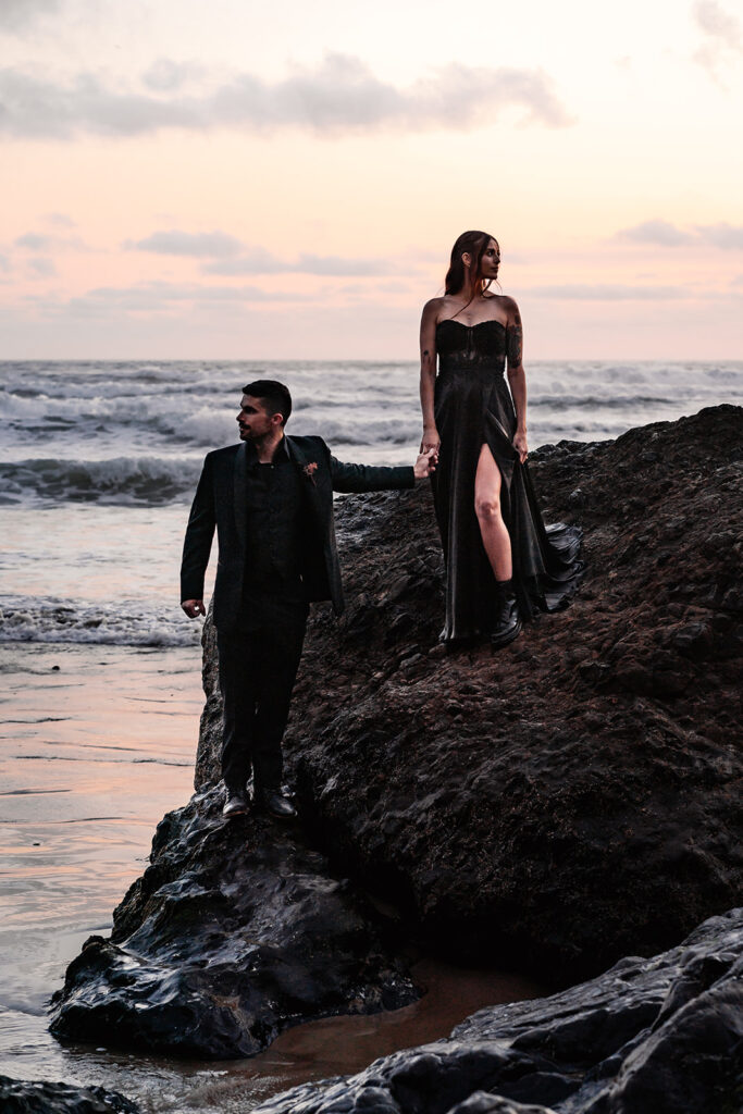 A bride and groom dressed in all black wedding attire, stand on a sea rock. The ocean waves churn behind them as they look in opposite directions