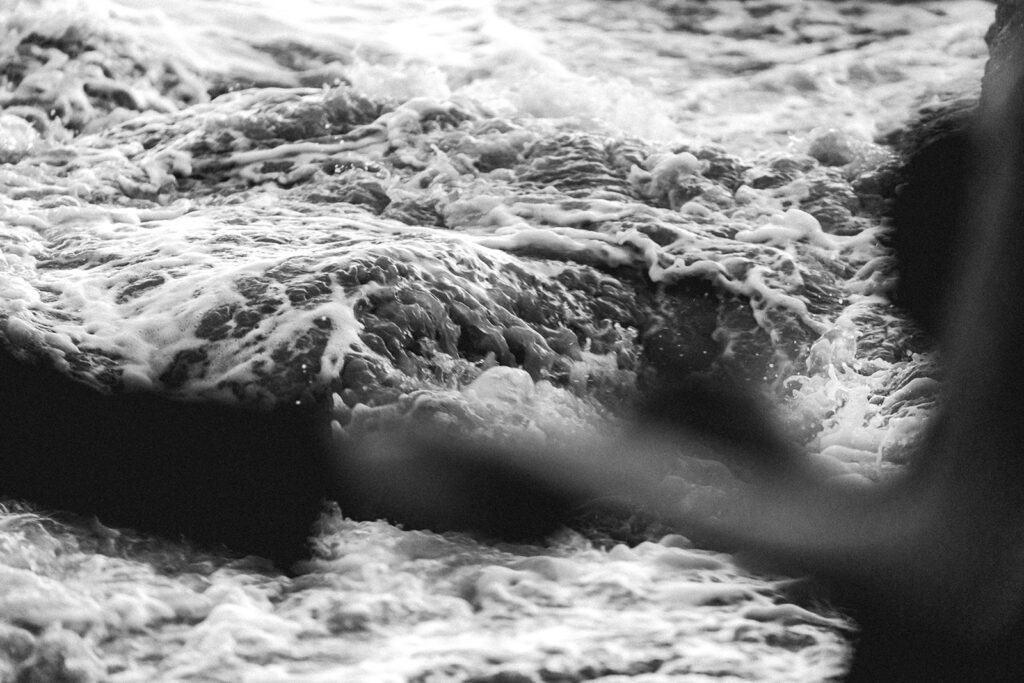 a bride and groom, exchange rings during their elopement ceremony. their hands are blurred as the shot focuses on the crashing waves behind them