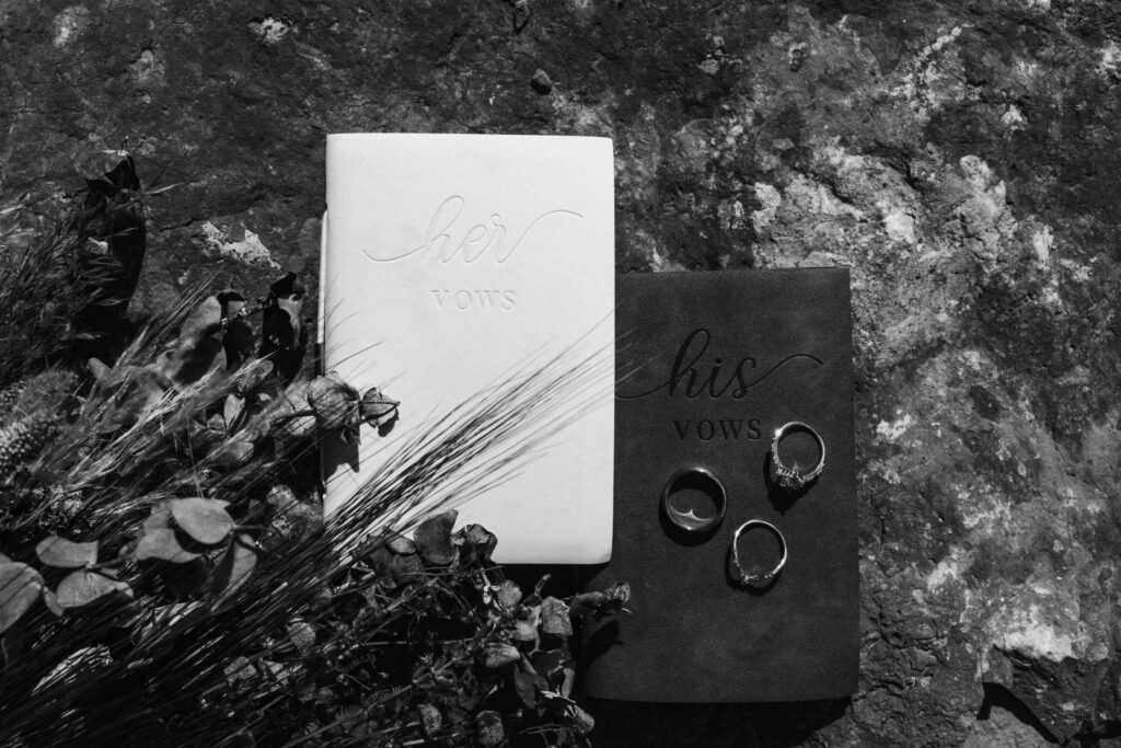 A wide, black and white image of our couples wedding details. We see their rings, vow books, and the bridal bouquet. 