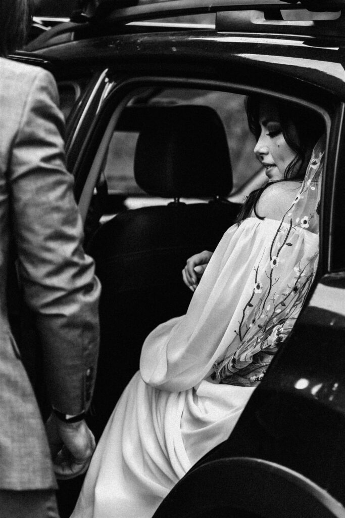 a black and wwhite photo of a bride in the car, her groom frames her as he helps her in to get ready for their sunrise elopement