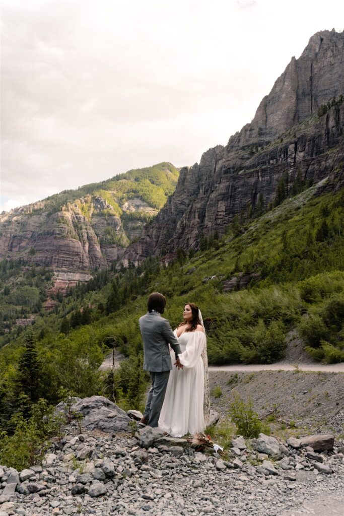 A couple in wedding attire stand on a rocky cliff. They gaze lovingly at each other as the sun rises behind them.