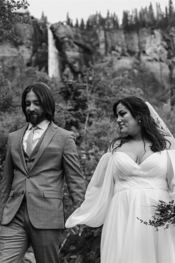 black and white image of a bride and groom waling hand in hand during their sunrise elopement. between them, in the distance, we see the tail of the falls they are headed to.