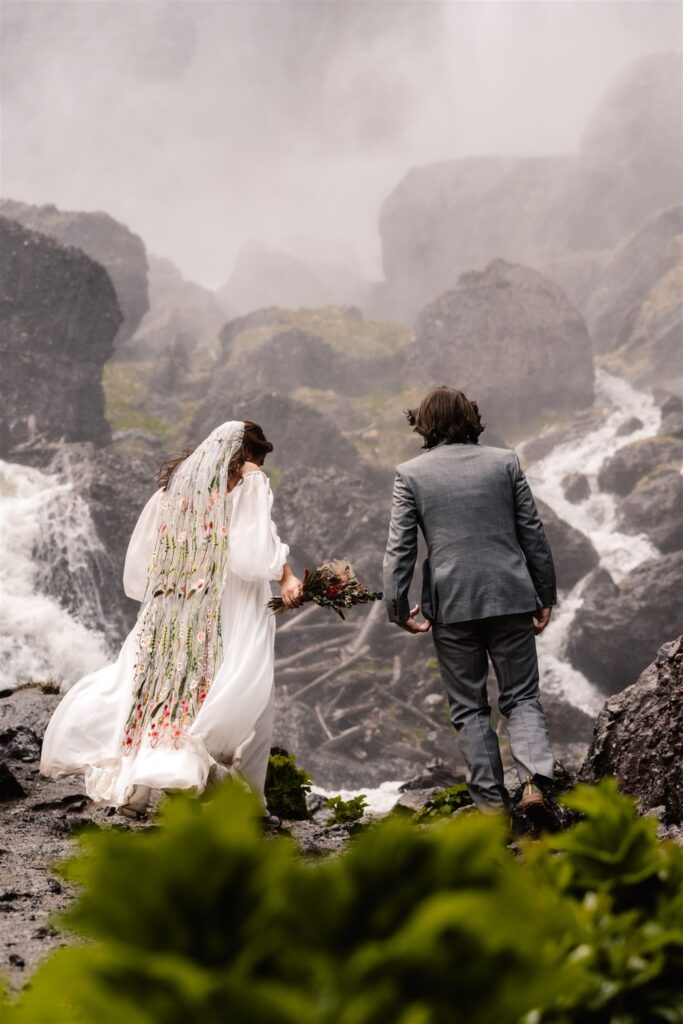 A couple in their wedding attire walk towards their ceremony spot during their sunrise elopement. You can see that the falls are mega powerful as the force of them makes our brides dress billow. They are surrounded by mist, moss, and large rocks.