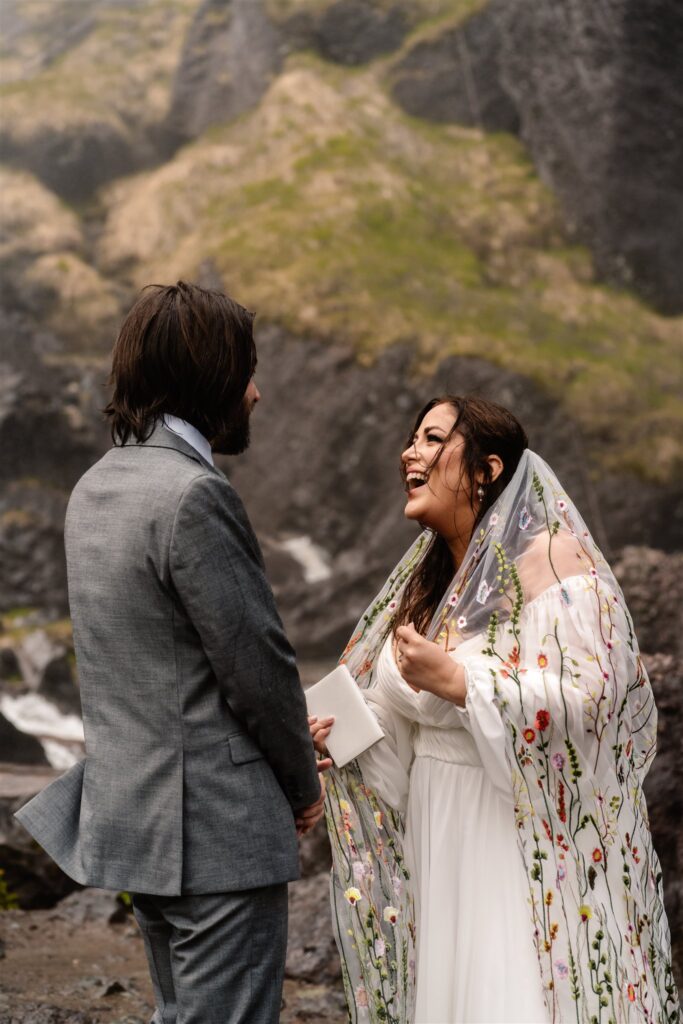 During their sunrise elopement ceremony a bride laughs and smiles widely as her groom reads his vows.