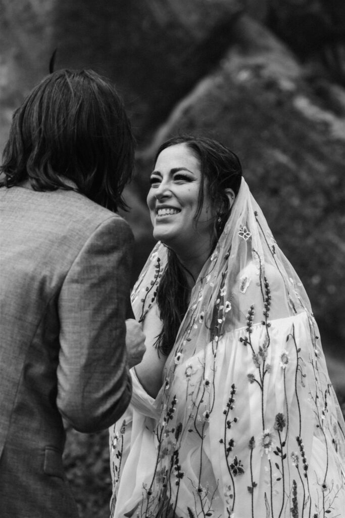 A black and white photo of a bride smiling widely as her groom reads his vows to her. Her veil, embroidered with wildflowers cascades over her shoulders. 