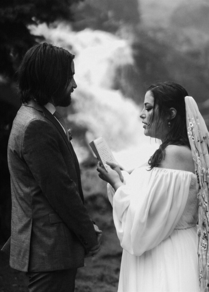 A black and white photo of a bride reading her vows to her groom. a waterfall gushes behind her