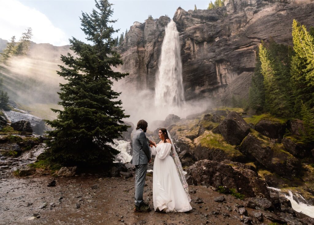 A landscape shot of their ceremony location for their sunrise elopement. A couple stands facing each other ith the waterfall gushing behind them. It creates a heavy mist that allows the morning light to filter through in the most stunning way.