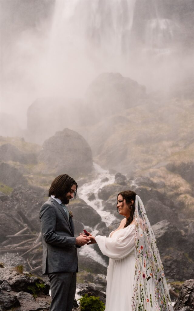 During their sunrise elopement, a bride and groom in wedding attire embrace in front of zig zagging falls. Moss covered rocks and waterfall mist make up the background. 