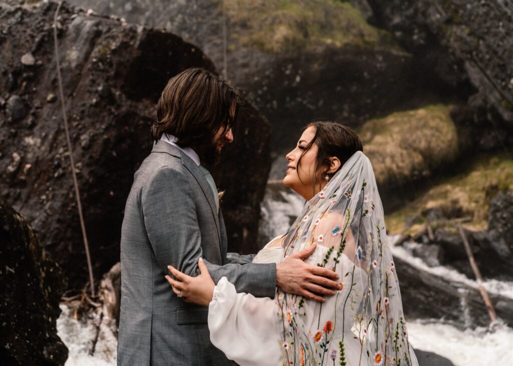 A bride and groom embrace during their sunrise elopement. Mossy rocks and waterfalls make up the background.